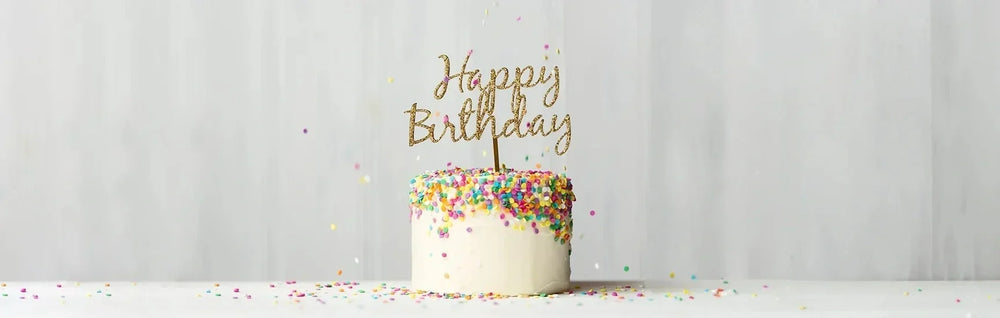 Birthday cake with colorful sprinkles and a glittery "Happy Birthday" topper against a soft neutral background.