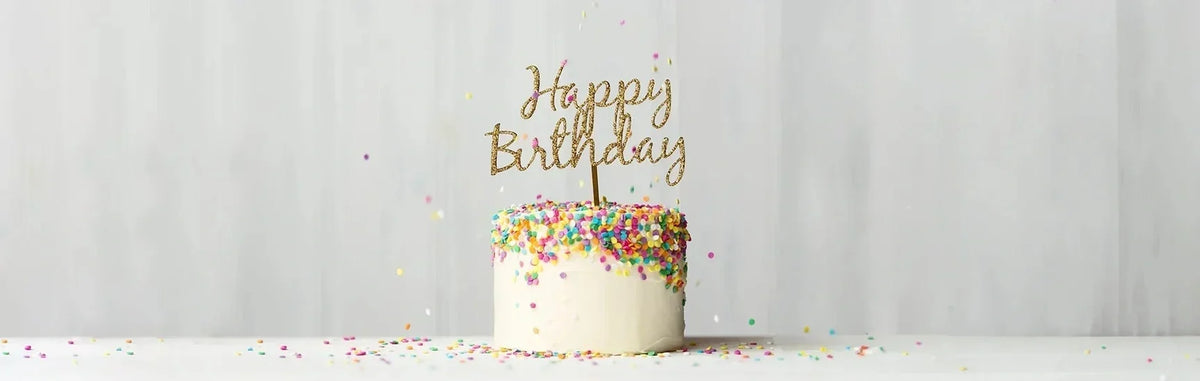 Birthday cake with colorful sprinkles and a glittery "Happy Birthday" topper against a soft neutral background.