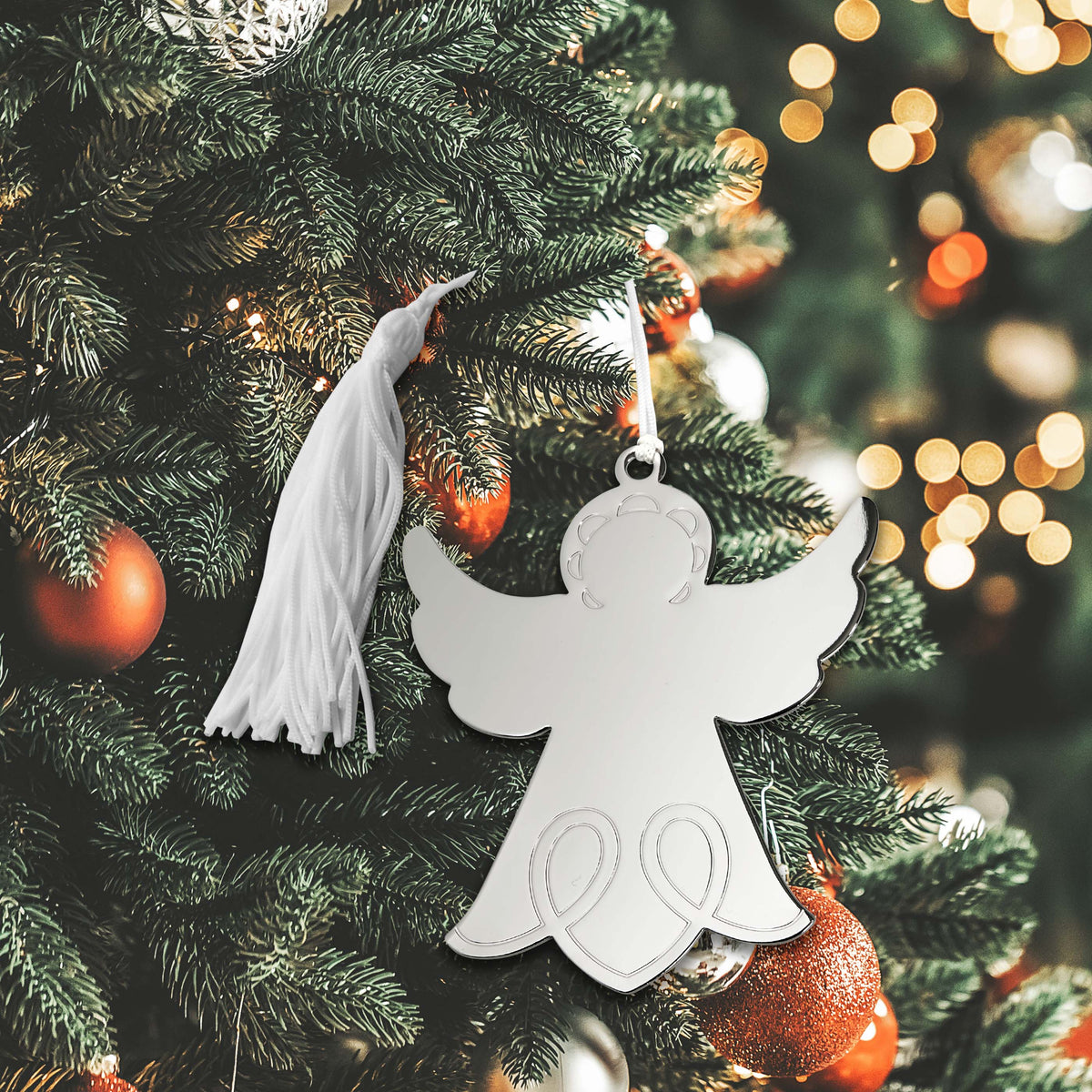 A bright, nickel-plated angel ornament with a white tassel hangs on a Christmas tree with festive red and gold baubles.