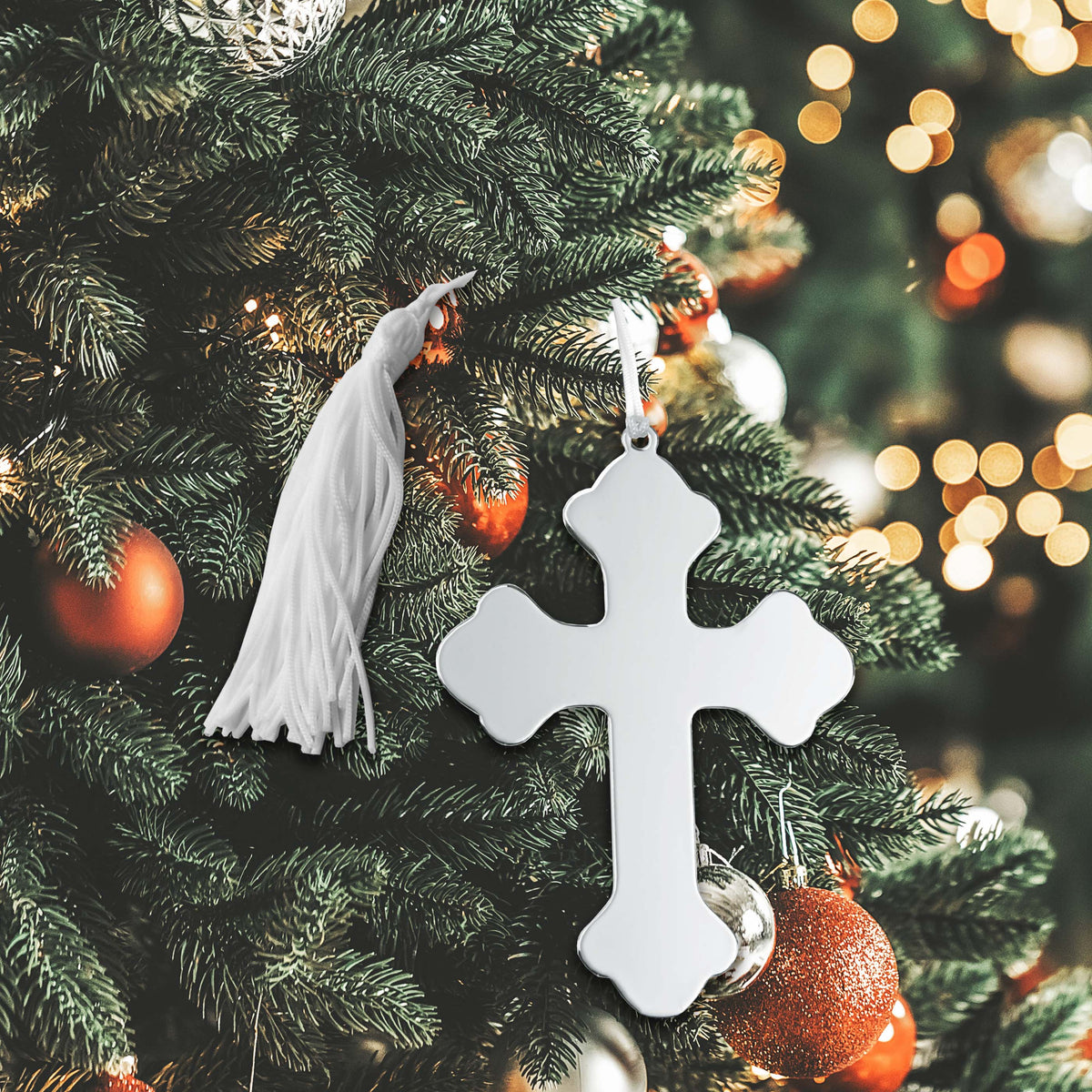 A silver cross ornament with a white tassel hangs on a Christmas tree with red and gold baubles.